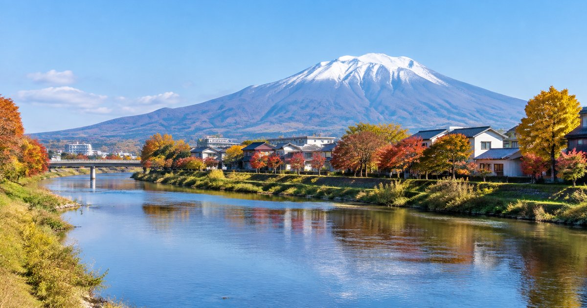 秋の川沿いの家並みと紅葉、遠くに雪をかぶった山が見える岩手の町